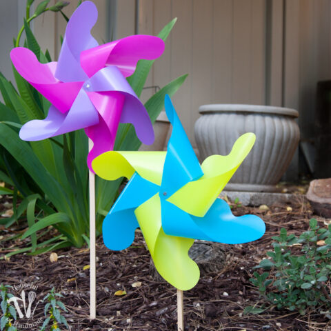 Two DIY giant outdoor pinwheels, one with pink and purple vanes and the other with blue and green, stuck in a flower garden.