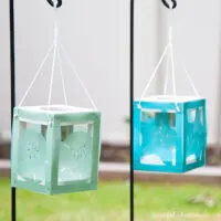 Two coastal themed wood lanterns hanging on a hook in the yard with solar lanterns inside.