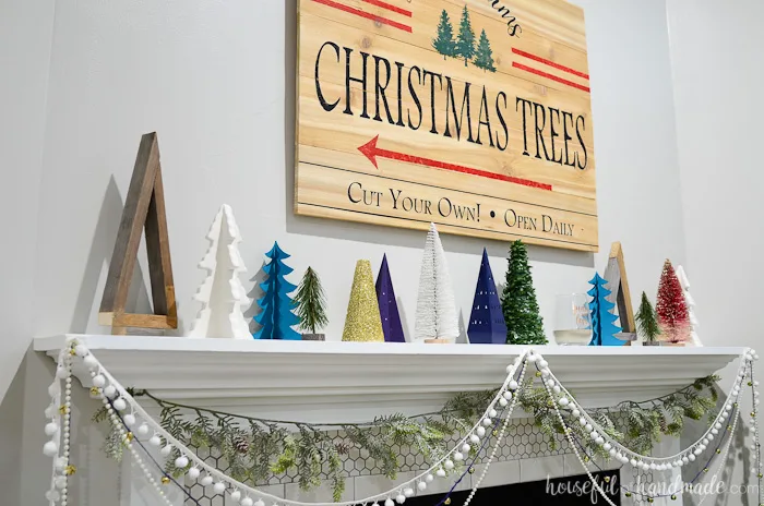 Decorative Christmas trees in non-traditional Christmas colors decorating a holiday mantel with a Christmas tree farm sign made from cedar fence pickets behind them.