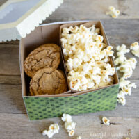 Green dotted paper cookie box with 2 compartments inside, one filled with cookies and the other with popcorn, with the lid sitting on the egde.