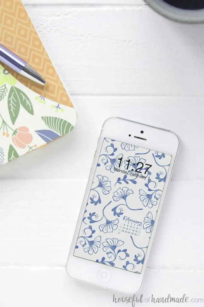 Smartphone with the blue floral digital background on the home screen on a desk next to a notebook. 