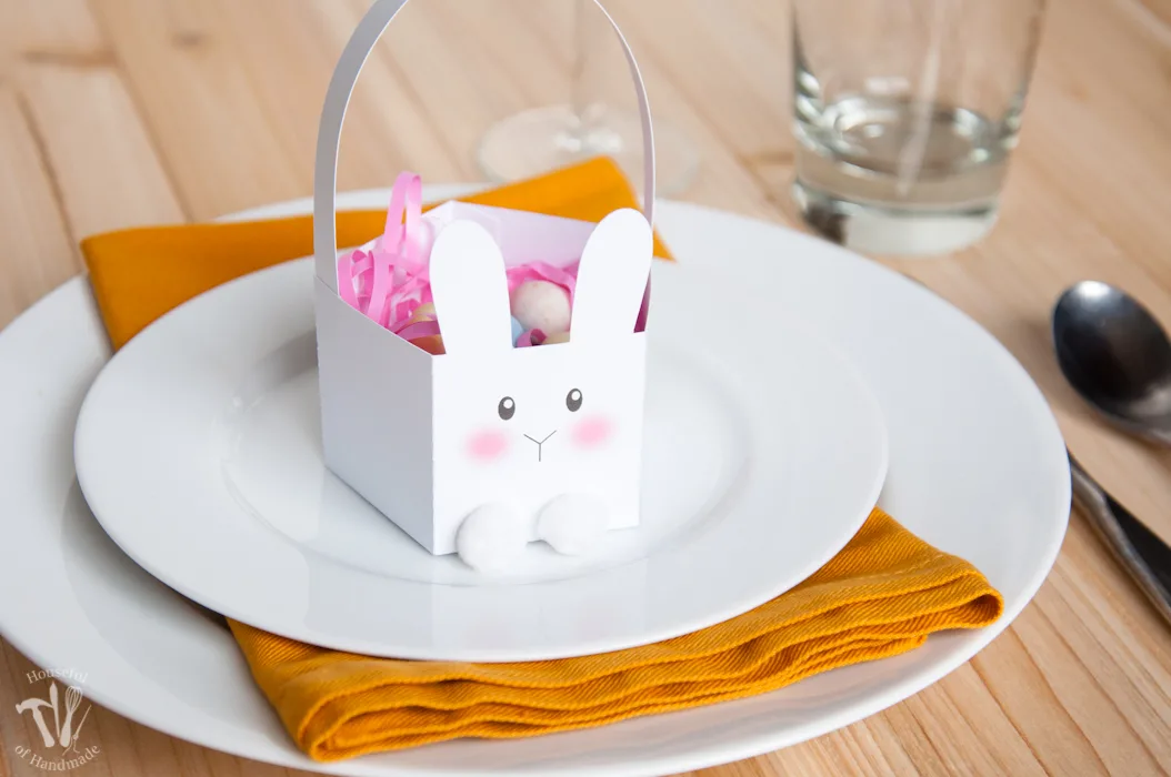 Easter table set with white plates and yellow napkins with a paper Bunny basket holding candies on top. 