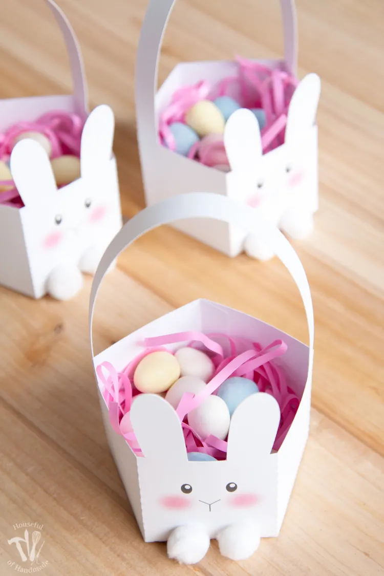Three white bunny Easter baskets with pink Easter grass and easter egg candies inside. 