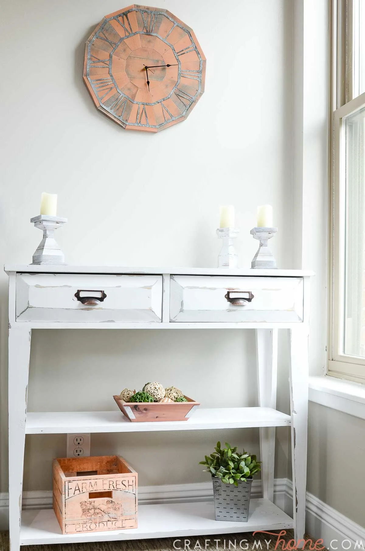 Small white console table with farmhouse decor on it and the paper wall clock hanging above it. 