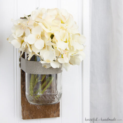 White flowers in a glass wall vase with reclaimed wood backing hanging on a wall.