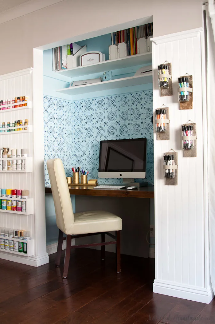 Desk in a closet with stenciled focal wall and wall vases on one side holding colorful markers and paint storage on the other side. 