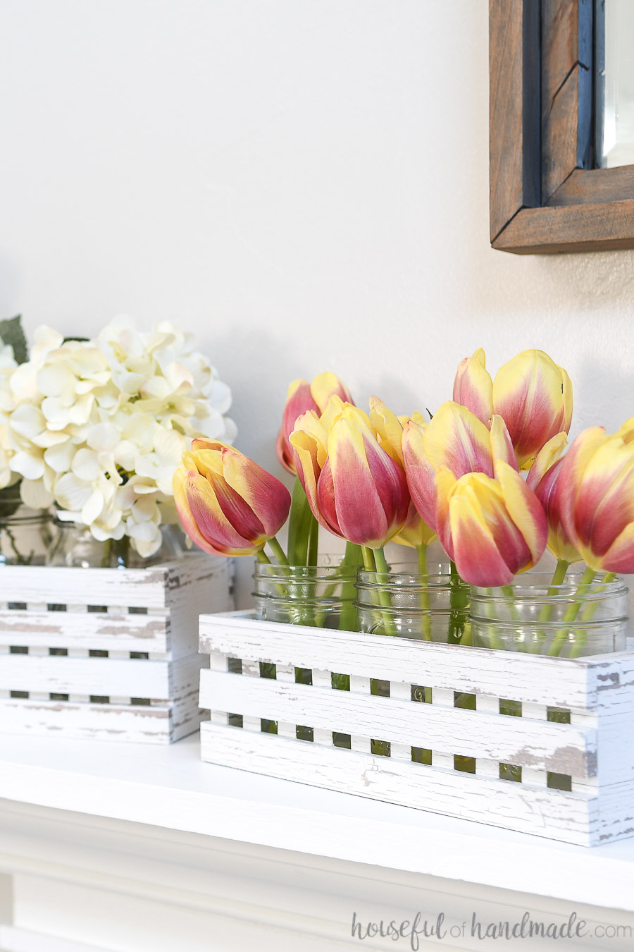 Two white chippy paint trough boxes on the mantel with mason jars filled with flowers inside. 