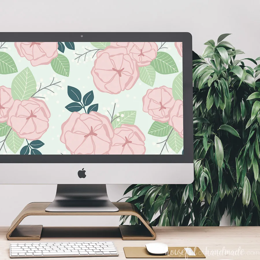 iMac on a desk in front of a plant with a pink and green floral wallpaper on the background.
