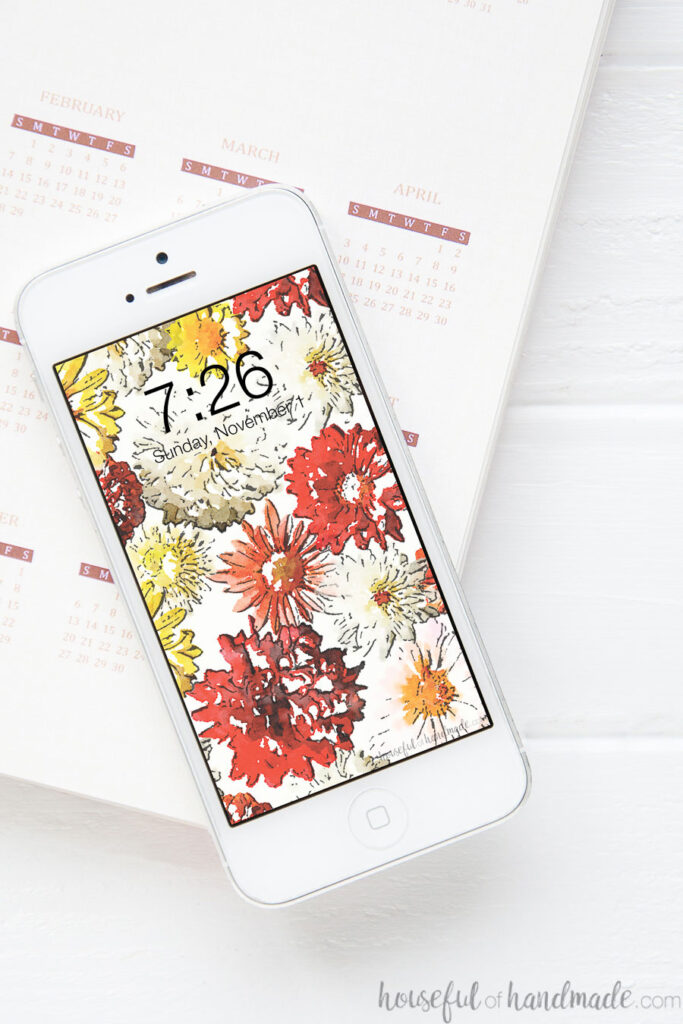 Smartphone sitting on an open planner page with a fall mums digital wallpaper on the screen.