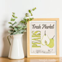 White vase with eucalyptus leaves next to maple frames with fall printable art in it, designed around a watercolor pear.