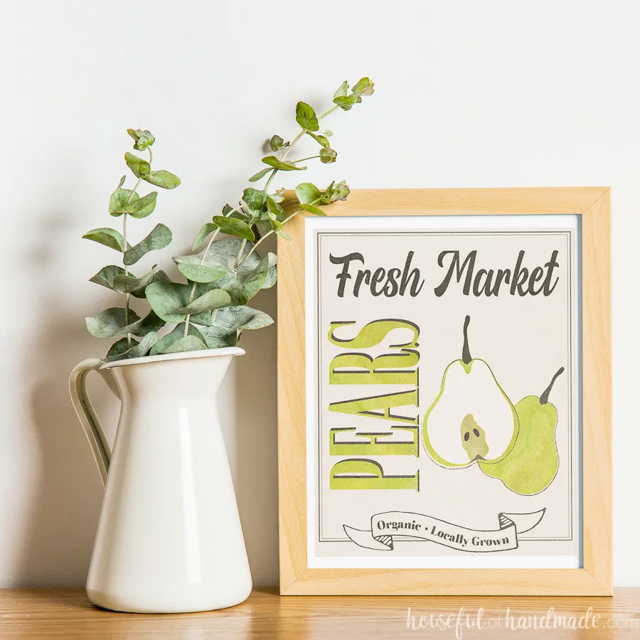 White vase with eucalyptus leaves next to maple frames with fall printable art in it, designed around a watercolor pear. 