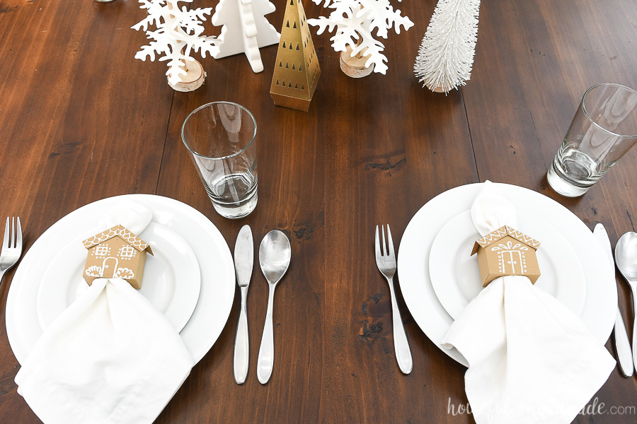 Two place settings on the side of the table with the gingerbread house napkin rings sitting on the place settings. 