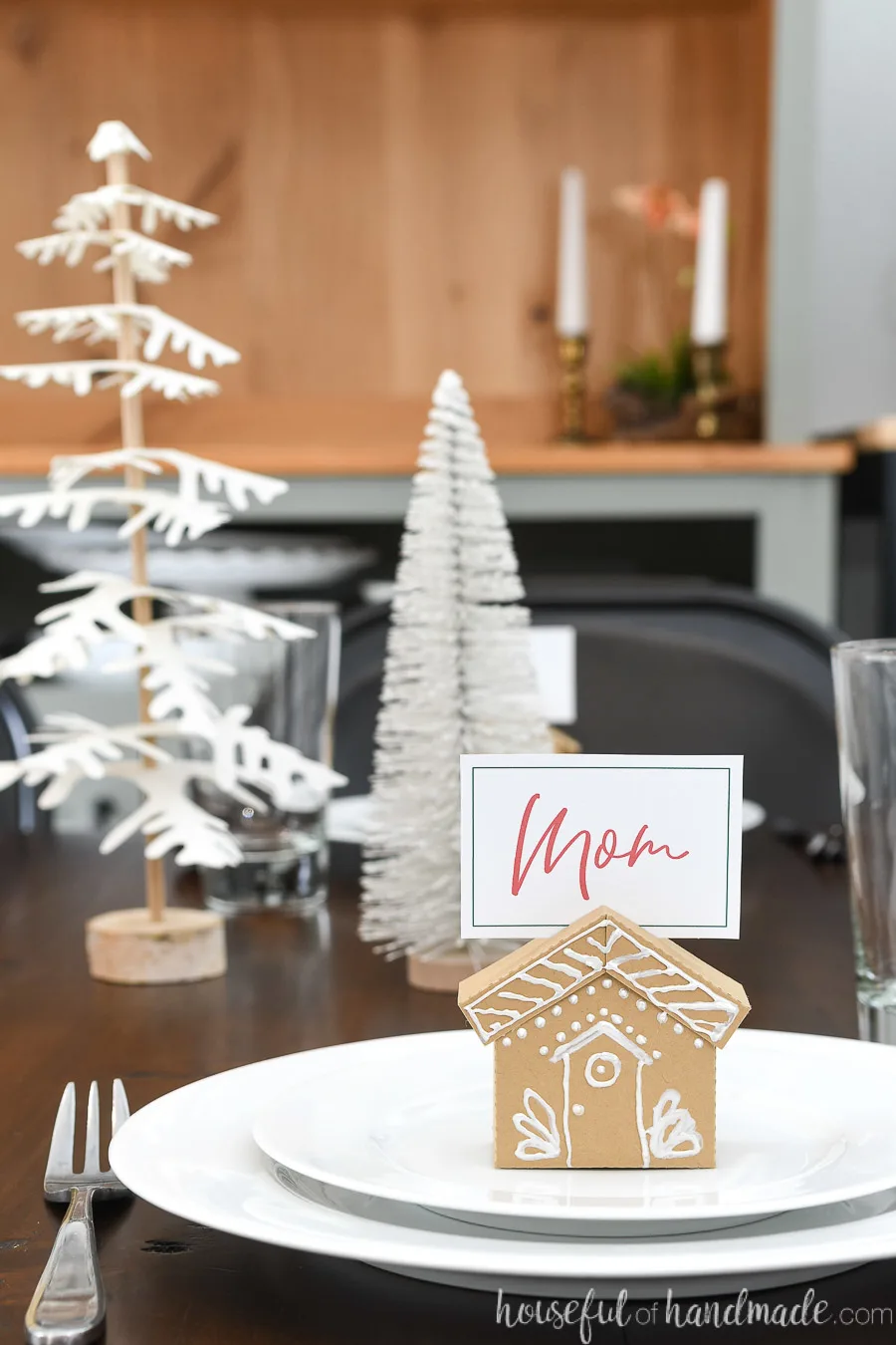 Christmas table set with Christmas trees as the centerpiece, white plates, with a brown paper gingerbread house place card holder sitting on them.