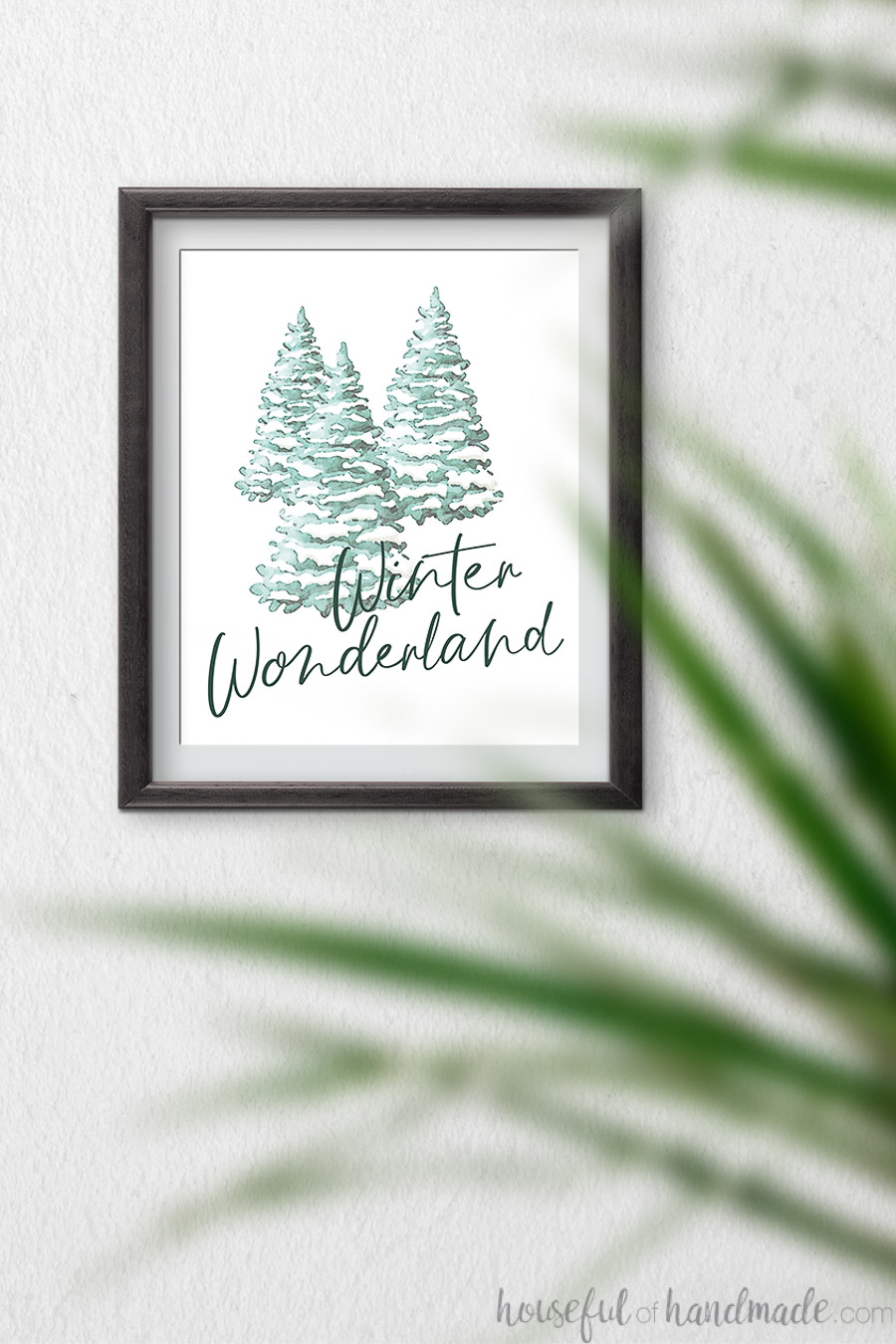 Watercolor Christmas printable with snow covered trees and script writing of Winter Wonderland on it, hanging on a wall.