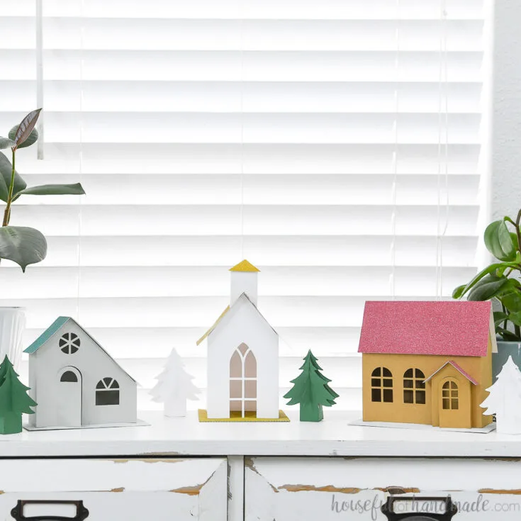 Putz inspired paper Christmas village with 3 different houses and paper Christmas trees on a console table.