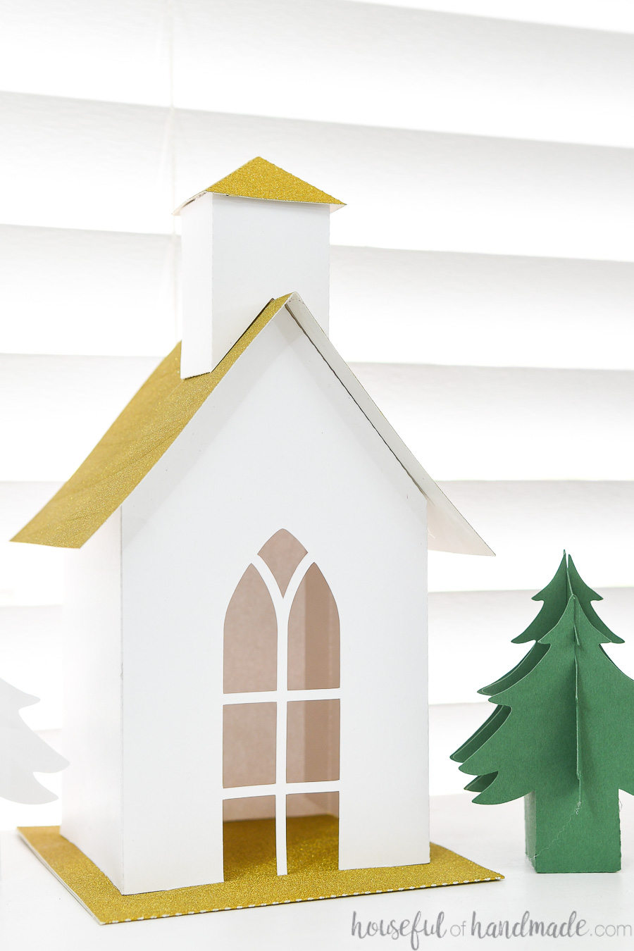 Close up of the paper Christmas church made with white cardstock and gold glitter roof and base next to a green paper Christmas tree. 