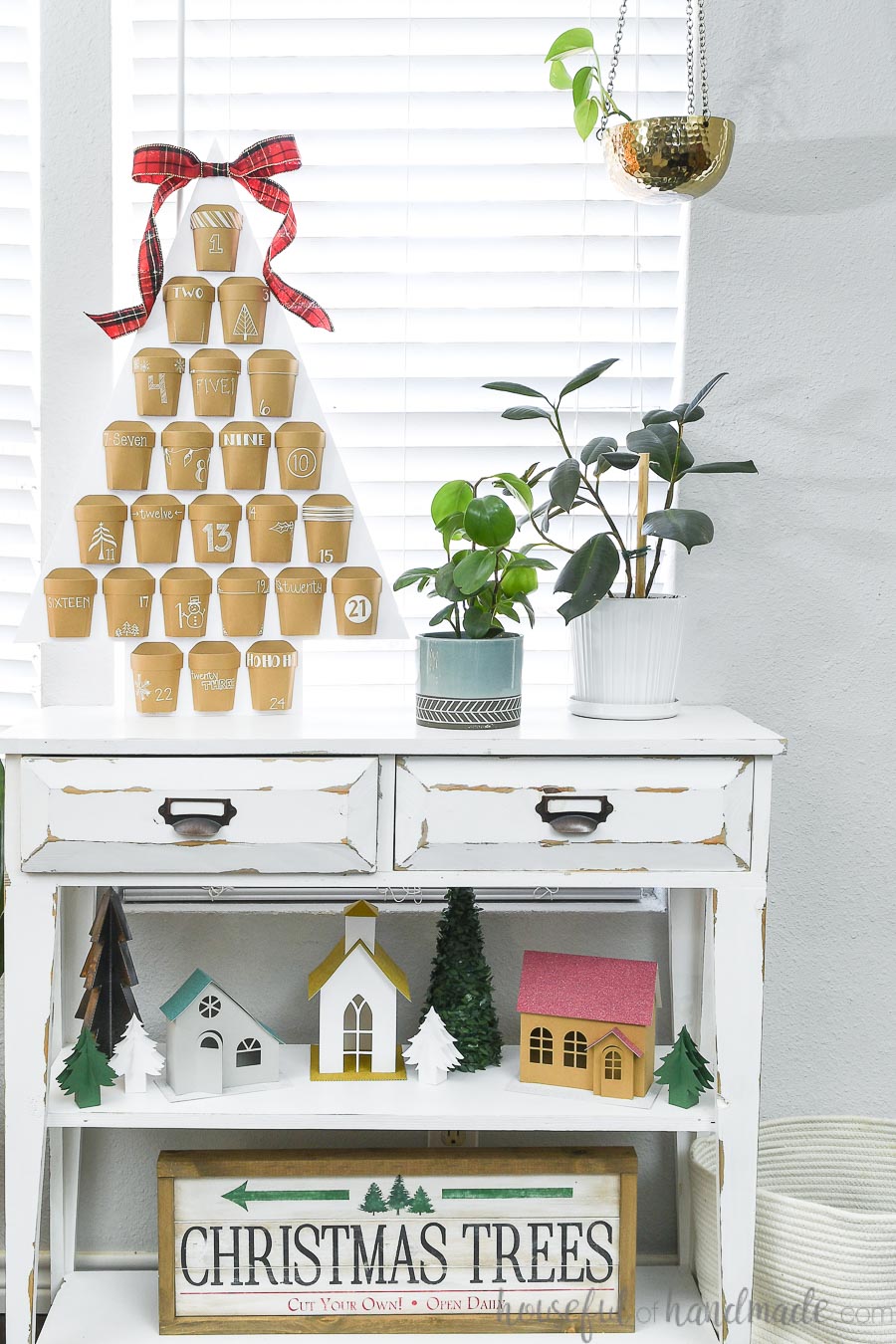 Christmas vignette with a Christmas advent calendar sitting on top of a white console table, a paper Christmas village on the middle shelf and a Christmas tree farm sign on the bottom shelf.