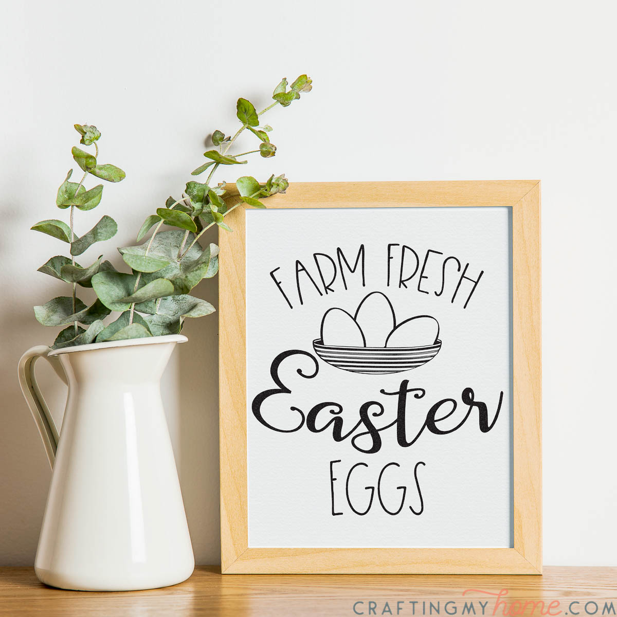 White pitcher with eucalyptus leaves in it next to a natural wood framed beautiful printable Easter sign.