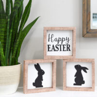 Three simple Easter signs stacked on a mantel next to a snake plant.