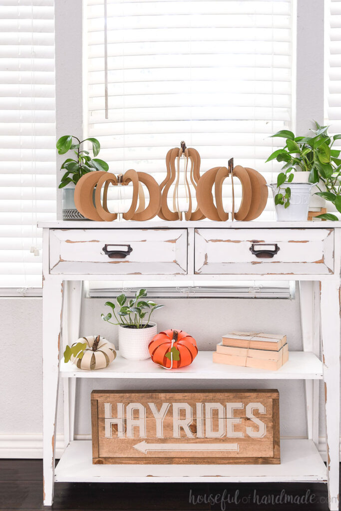 White farmhouse console table decorated for fall with 3D pumpkin lanterns, paper pumpkins, plants, books, and a wooden hayrides sign.