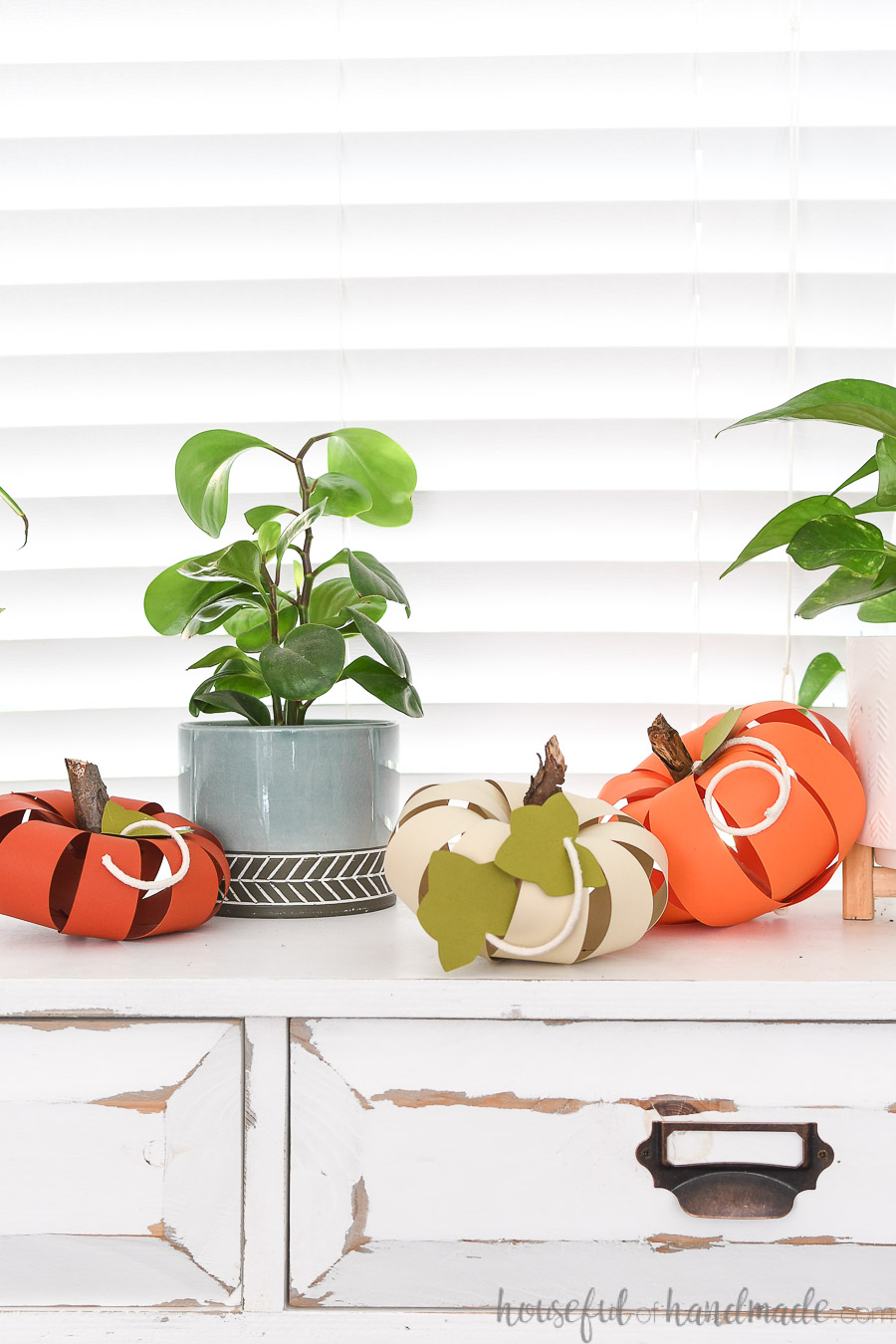 Console table with 3 paper pumpkins in oranges and cream next to two potted house plants.