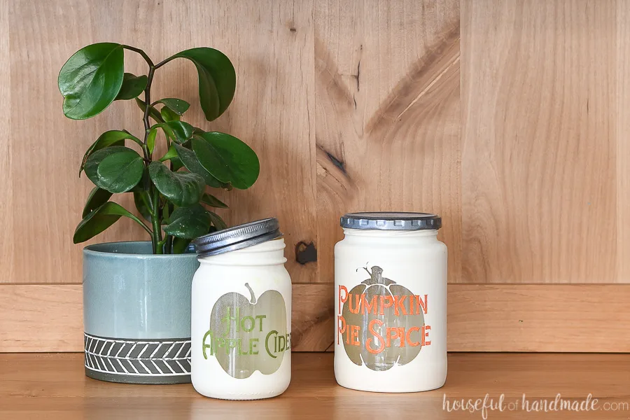Two fall themed kitchen canisters in a wood hutch with a houseplant in the background.