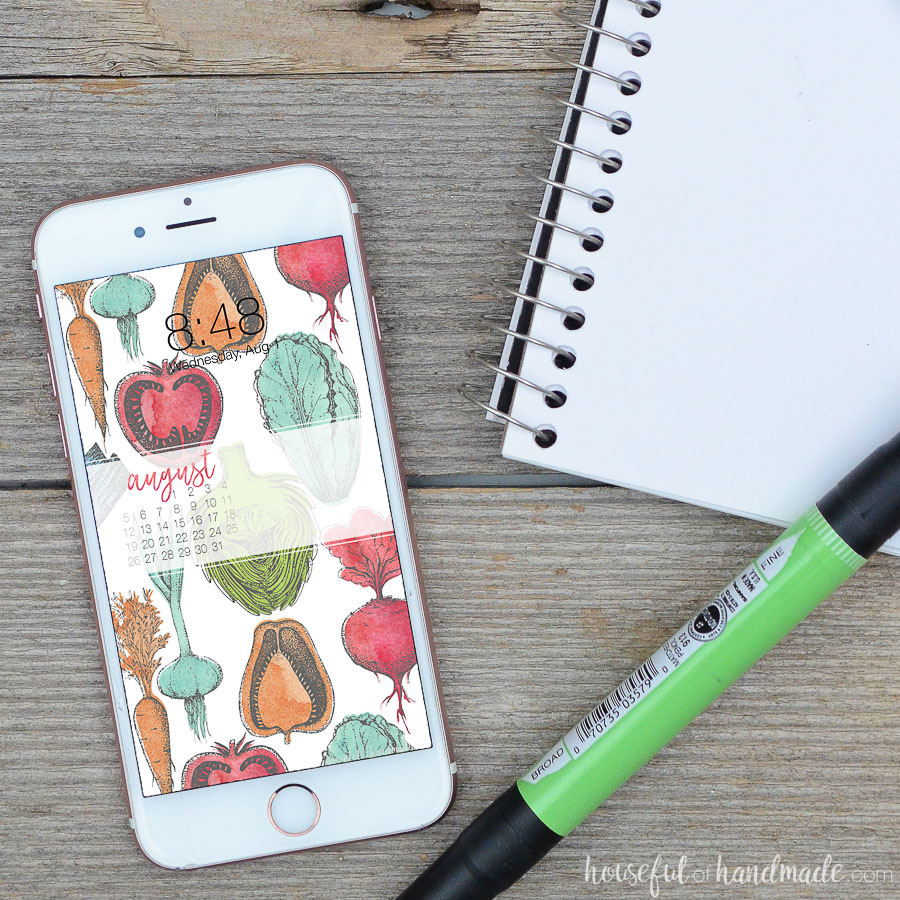Digital background with August calendar. Free digital wallpaper of colorful hand-drawn vegetables on a smartphone screen next to sketchbook. 