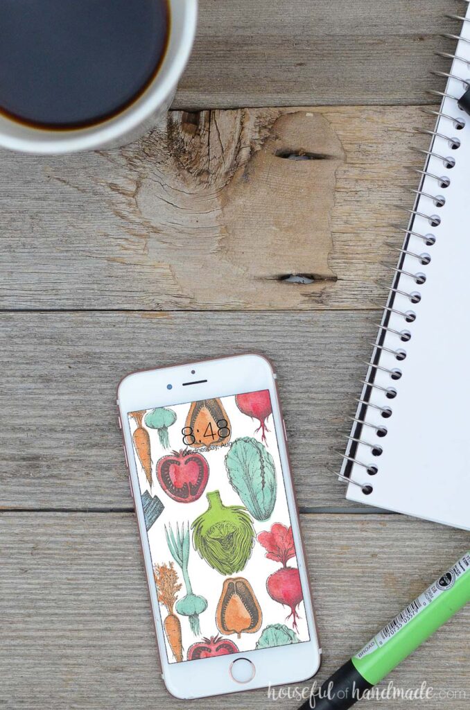 White iPhone smartphone with colorful vegetable design digital wallpaper on it. Phone is laying on a wood background with sketchbook and cup of coffee next to it. 