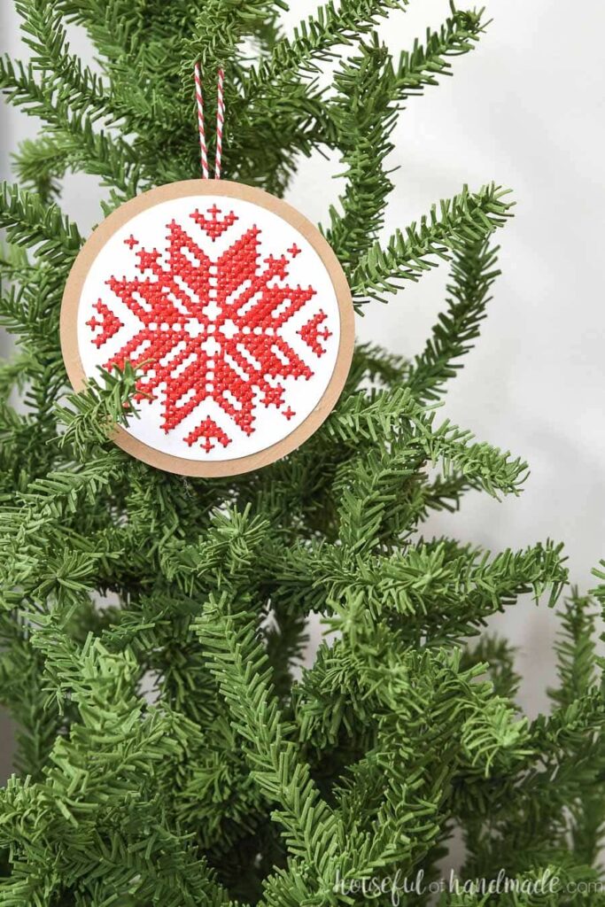 Round cross-stitched paper ornament with Nordic snowflake design hanging on a tree.
