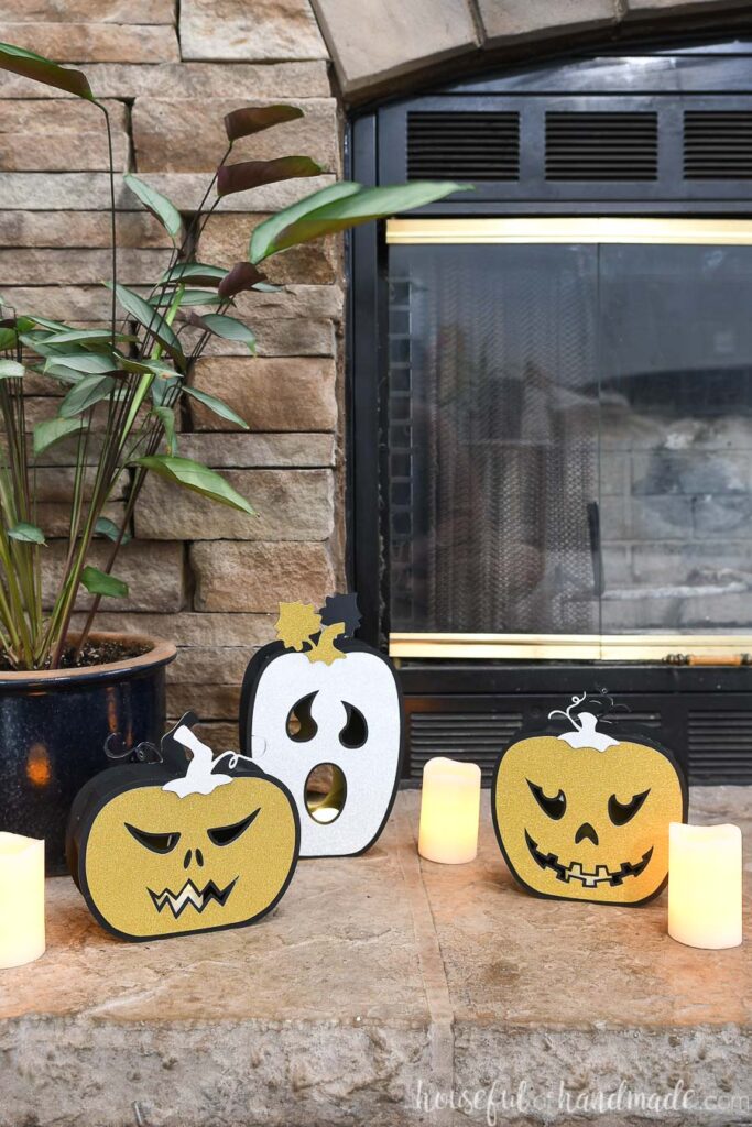 Two standard jack-o-lantern faced and one ghost faced paper jack-o-lanterns on the hearth with candles next to them. 