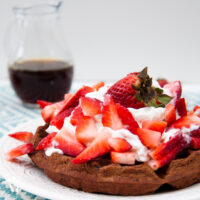 Healthy chocolate waffles on a white plate topped with whipped cream and fresh strawberries.