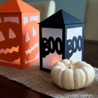 Black boo lantern next to white mini pumpkin with orange pumpkin lantern in the background.