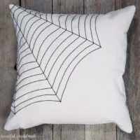 Drop cloth throw pillow with spiderweb design sewn in one of the corners laying on a wood background.