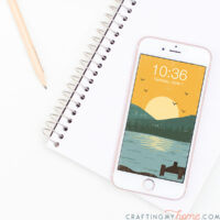 White iPhone with a vintage inspired lake sunset digital wallpaper on the home screen sitting on a sketchpad.