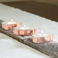 Three copper candle holders in the shapes of fall leaves and acorns on a rustic board.