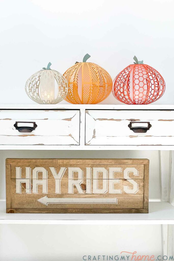 White farmhouse console table with cream, yellow-orange, and orange paper pumpkin lanterns on it along with a Hayrides wood sign. 