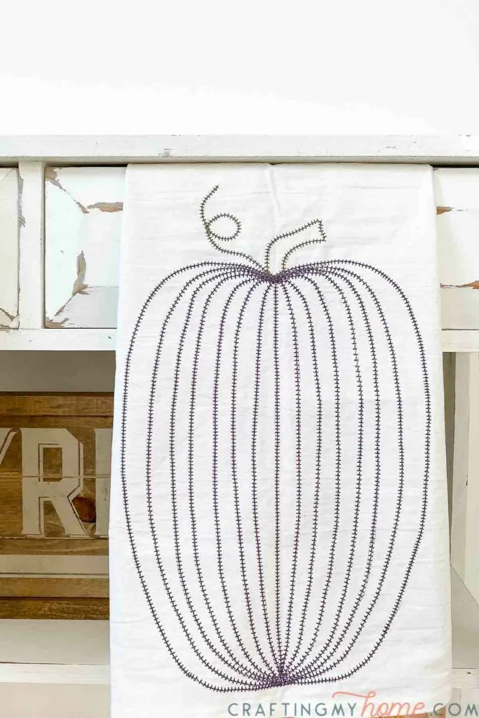 Tall pumpkin design sewn into a tea towel with purple thread hanging on a console.  