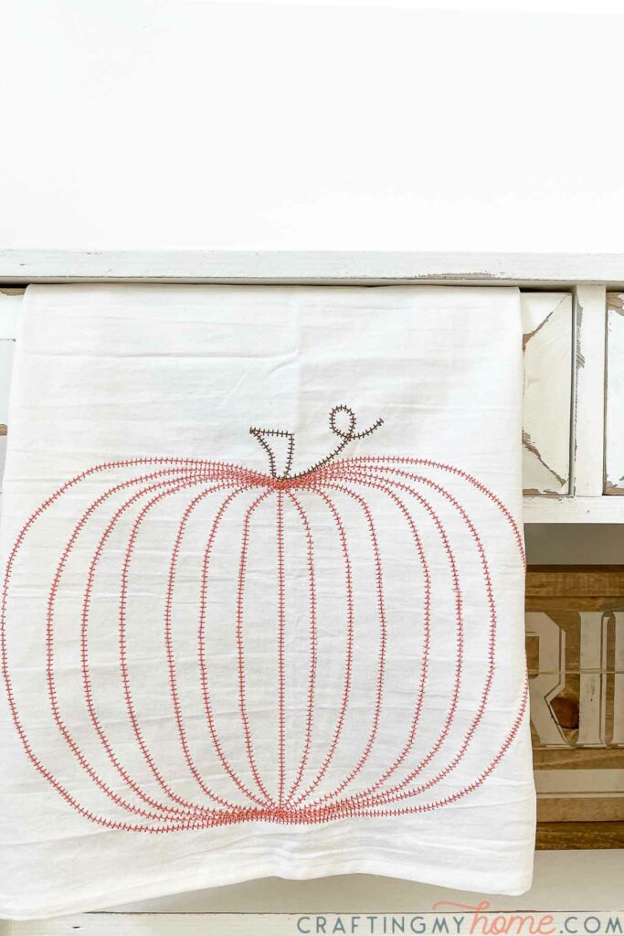 Round pumpkin design sewn into a tea towel hanging on a console table. 