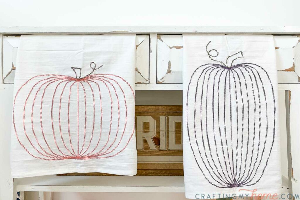 Two DIY tea towels with large pumpkins stitched into them hanging on a console table. 