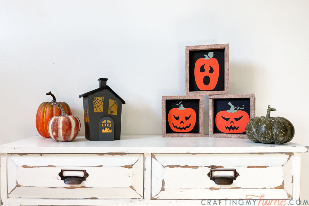 Halloween vignette on a console table with a haunted house halloween lantern, fake pumpkins, and three wood framed signs with jack-o-lantern faces on them.
