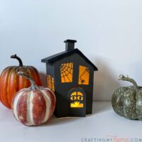 Haunted house halloween lantern made from paper with flameless tea light inside and pumpkins around it.