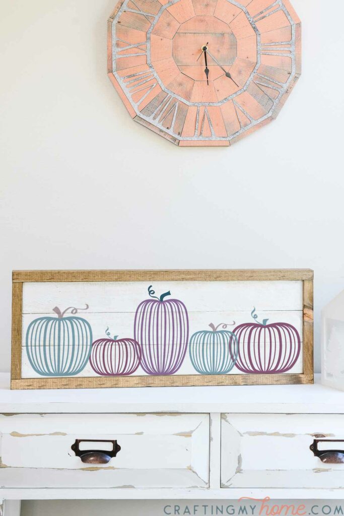 Horizontal wood sign with five modern pumpkin SVG files cut out of vinyl on it, sitting on a console table below a clock. 