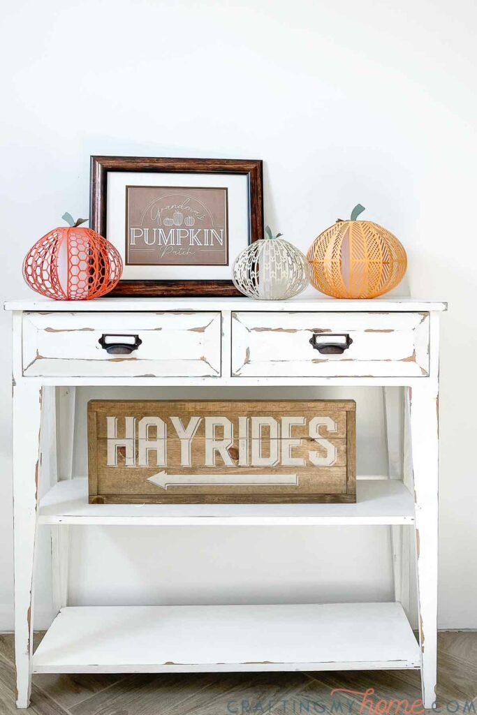 White farmhouse console table decorated for fall with a neutral pumpkin patch printable sign, paper pumpkin lanterns, and a wooden hayride sign. 
