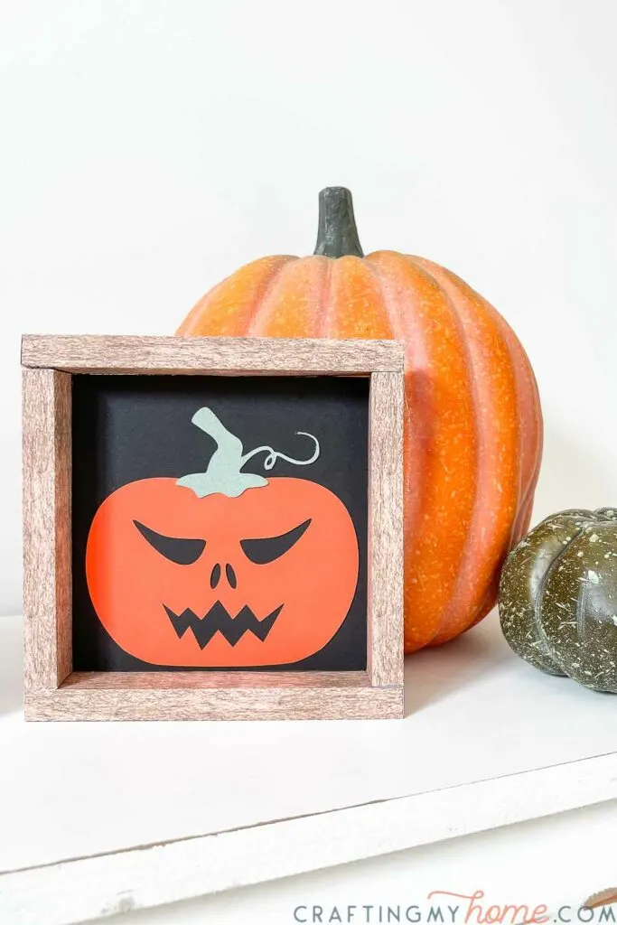 Spooky jack-o-lantern design on a faux wood framed sign in front of a pumpkin. 