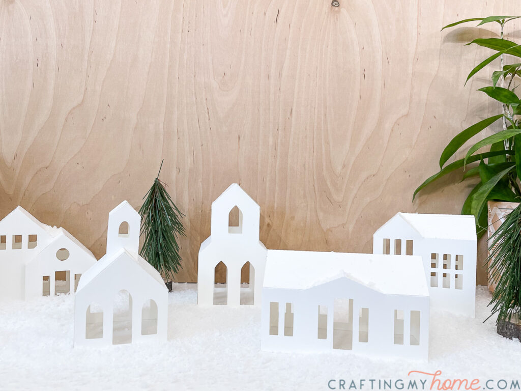 Five white paper Christmas houses on a table surrounded by fake snow with some greenery.