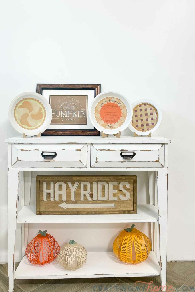 White console table decorated for Thanksgiving with DIY decorative pie plates, paper pumpkin lanterns, Hayrides sign, and pumpkin patch art. 