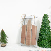 Decorative wooden sled on a table covered with fake snow next to decorative trees.