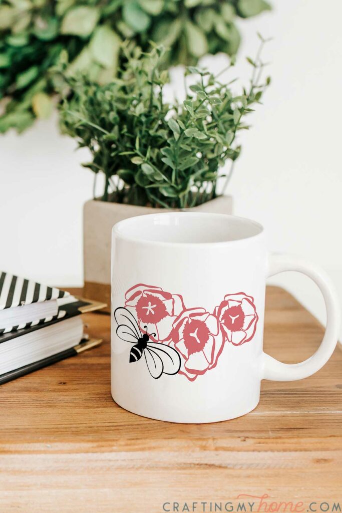 Three flowers and a bee cut file on a white mug sitting by greenery. 