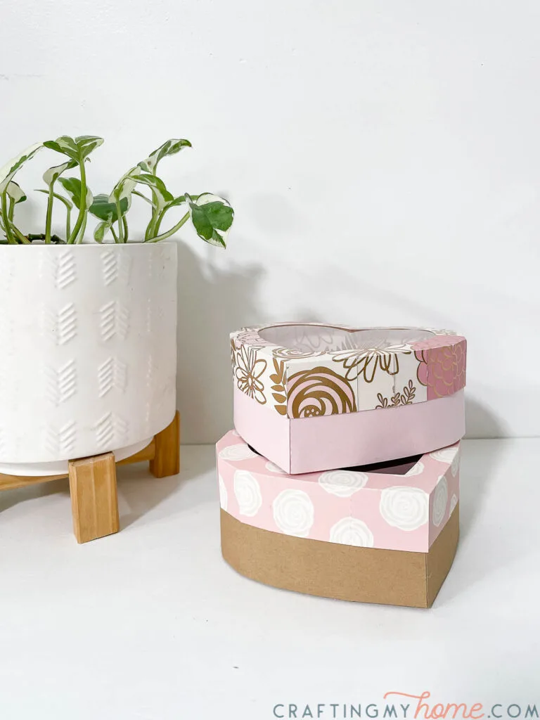Two heart gift boxes with floral patterned lids stacked next to a plant. 