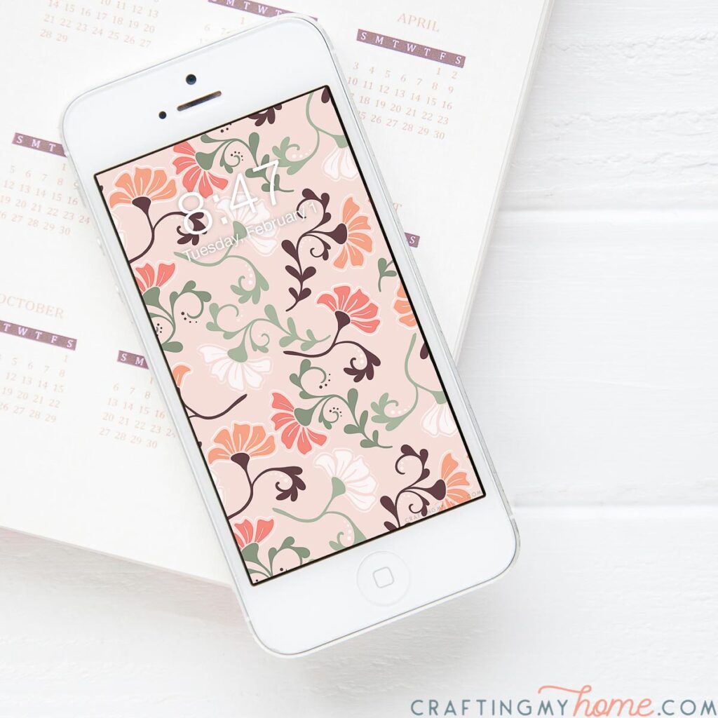 Smartphone showing the pink floral digital wallpaper on the home screen sitting on a planner. 